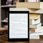 une pile de livres sur une table en bois. devant eux est posée une liseuse électronique allumée sur une page de livre.